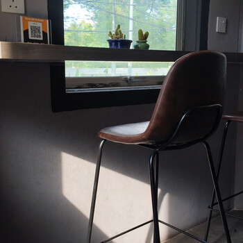 Modern bar stool with brown leather seat and black metal frame positioned at a counter near a window with plants on the shelf.