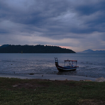 Wooden fishing boat moored in shallow water at dusk, with forested shoreline and mountains in the distance.