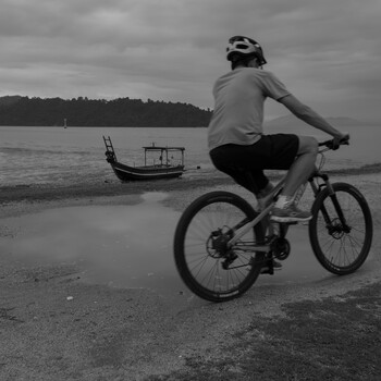 Cyclist in helmet and casual clothing riding on beach sand with traditional longtail boat anchored in calm water behind.