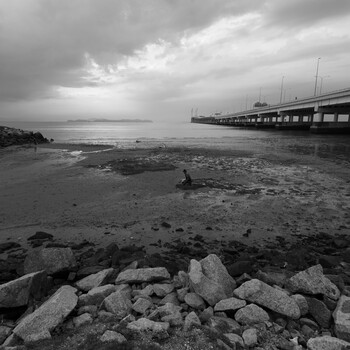Rocky foreground leads to a person standing in shallow water, with a pier extending into calm bay under dramatic storm clouds.