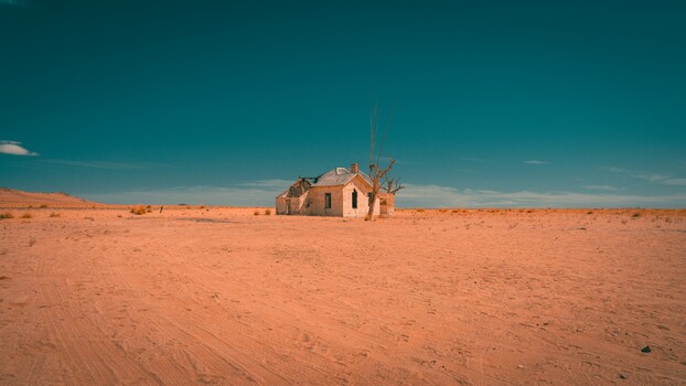 Abandoned brick house with deteriorating walls standing alone in a vast, flat desert under clear blue sky.
