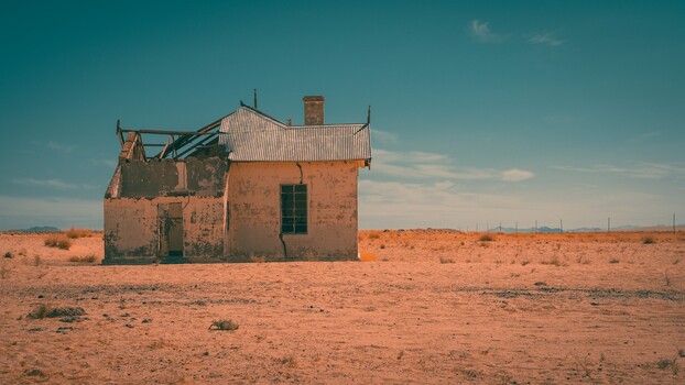Abandoned brick house with corrugated metal roof in vast desert landscape under clear blue sky.