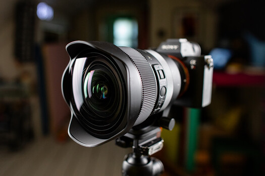 Wide-angle lens mounted on mirrorless camera body with hood attached, positioned on tripod in indoor setting.