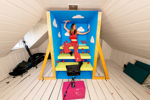 Overhead view of a woman in red checkered outfit posing on yellow and blue striped deck chair set against painted blue sky backdrop.