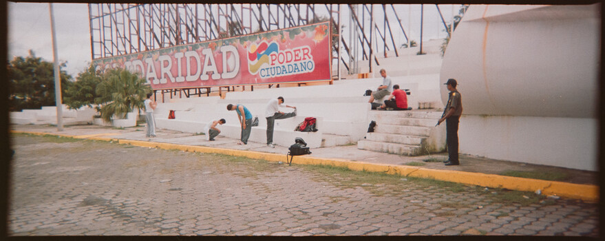 People sitting on white concrete steps in a public plaza with a large political banner overhead and yellow-painted curbs.