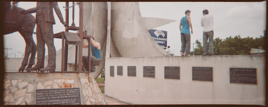 Monument with bronze statues and memorial plaques, two people walking on top of white structure.