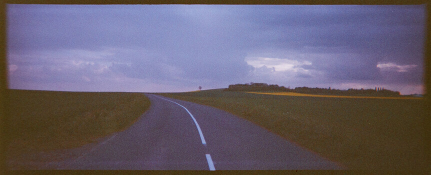 Straight asphalt road with white center line extending through flat agricultural fields under stormy purple sky.