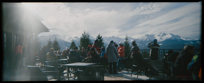 Crowd of people gathered on a sunny mountain terrace with snow-capped peaks in the distance.