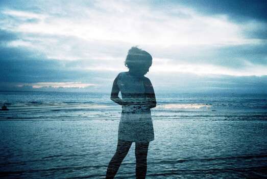 Woman standing in shallow ocean water at sunset, silhouetted against dramatic sky