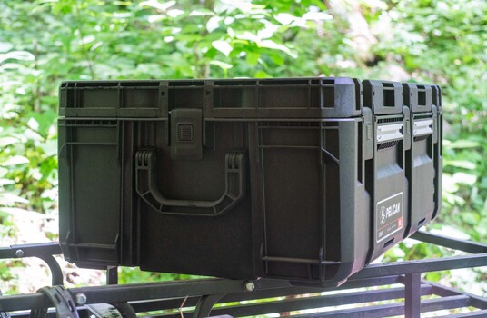 Black protective equipment cases stacked on outdoor metal shelving with green foliage background