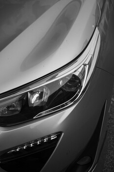 Close-up black and white photograph of a car's headlight and hood in strong side lighting
