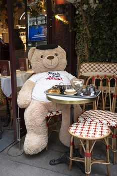 Large cream-colored teddy bear seated at a bistro table wearing a white t-shirt