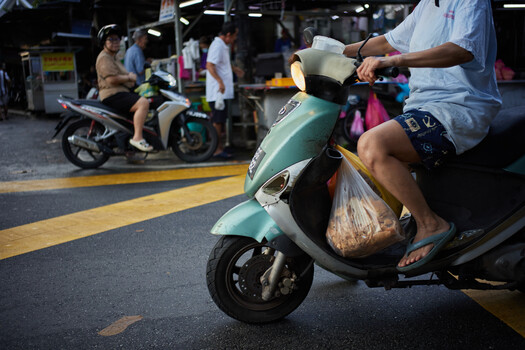 Man holding a plastic bag sits on a turquoise scooter at a street market, wearing a light blue shirt and hat