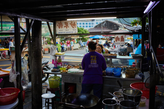 Street food vendor cooking in an open-air market stall with customers visible in the bustling background