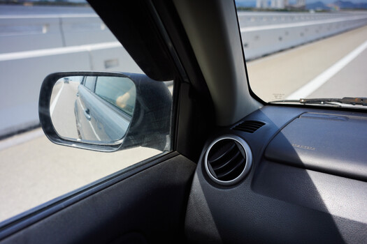 View from inside a car showing side mirror reflecting highway and dashboard air vent