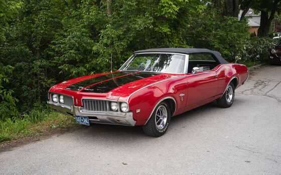 Red convertible classic car with hood open, parked against green foliage backdrop