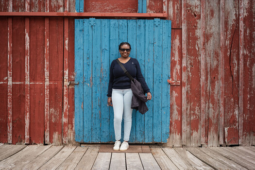 Woman in white pants and black top standing against weathered red wooden barn doors with bright blue central doors