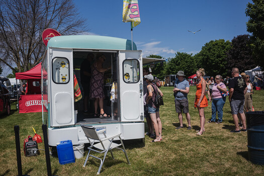 White food truck with open serving window surrounded by visitors at outdoor festival on sunny day