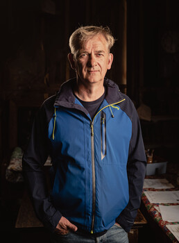 Middle-aged man in blue and black jacket smiling at camera against dark background