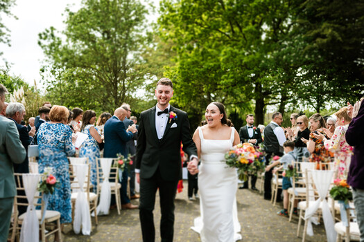 Bride and groom walk down outdoor aisle lined with seated guests and floral arrangements