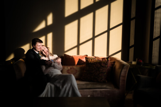 Man and woman in formal attire reclining on a dark leather sofa with dramatic window light creating geometric shadows across their bodies.