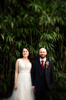 Bride in white lace dress and groom in navy suit with burgundy tie stand before dense green foliage
