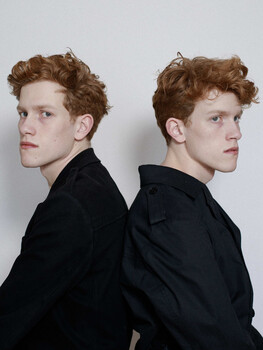 Two young men with reddish-brown curly hair shown in profile view wearing black shirts against neutral background