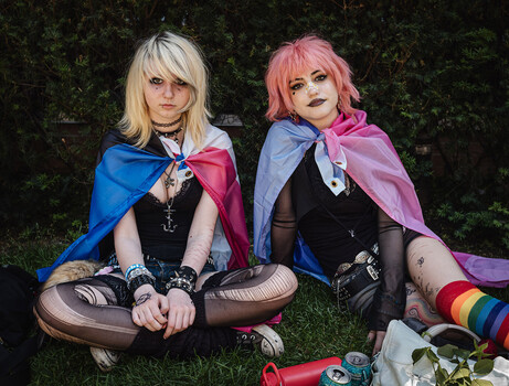 Two cosplayers sitting on grass wearing colorful capes and dark clothing against an ivy backdrop