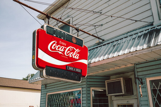 Vintage Coca-Cola neon sign hanging from a metal structure outside a turquoise-painted building