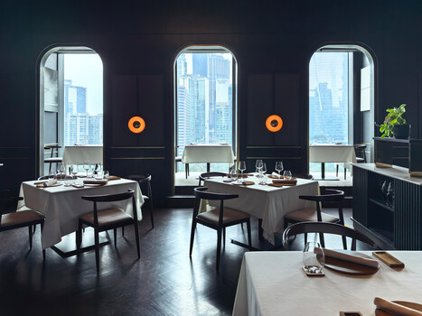 Modern restaurant interior with dark walls, floor-to-ceiling windows overlooking city skyline, and minimalist table settings