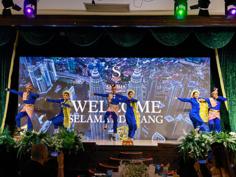 Stage production with dancers in blue and gold costumes performing before large projection screen displaying cityscape and welcome text