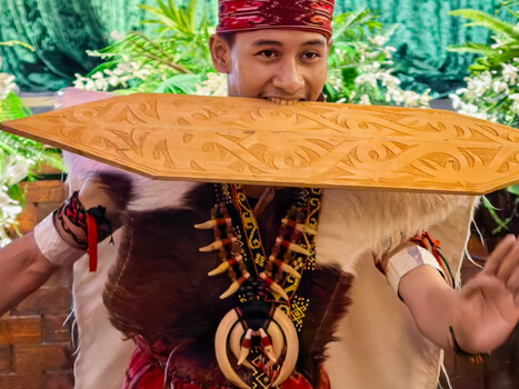 Young man holding ornately carved wooden shield with traditional red headwrap and patterned shirt