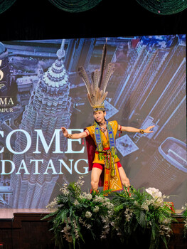 Performer in ornate gold and red Thai costume on stage with projected Bangkok cityscape backdrop