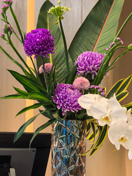 Purple pompom dahlias and white orchids arranged in a geometric blue vase with tropical foliage