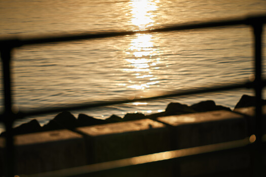 Sunset seascape framed through wooden railings, with golden light reflecting on calm water and silhouetted rocks