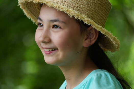 Young woman wearing straw hat and light blue shirt, smiling against soft green bokeh background