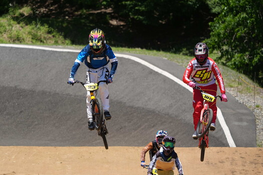 Three BMX riders racing on a dirt track with number bibs, captured mid-competition in bright daylight