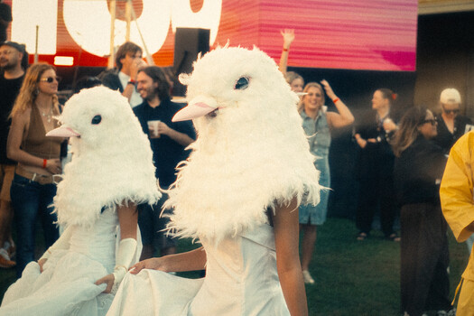 Two people wearing white feathered eagle head masks at an indoor event with spectators