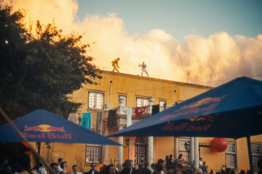 Two people balancing on the roof of a yellow building with dramatic clouds and mountains in background