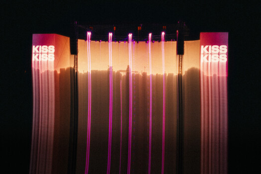 Neon installation with vertical tubes in pink, magenta, and warm tones against dark background with 'KISS' text signage