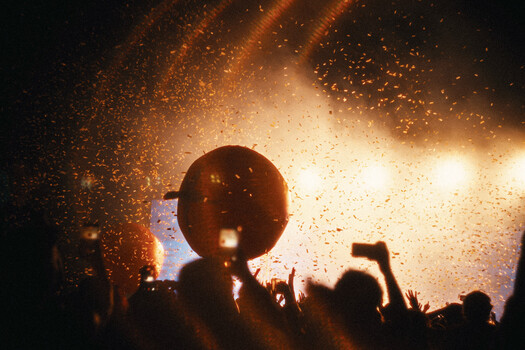 Silhouetted crowd with raised phones backlit by intense golden stage lighting and pyrotechnics