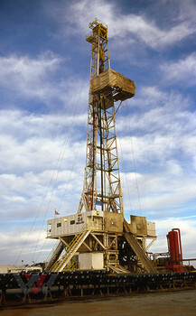 Tall yellow oil derrick under dramatic cloudy sky with visible substructure and equipment