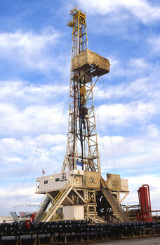 Tall oil derrick drilling rig with yellow structural framework against blue sky with white clouds