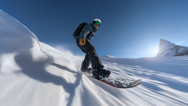 Skier in dark jacket carving down steep snowy slope with dramatic mountain backdrop and clear blue sky