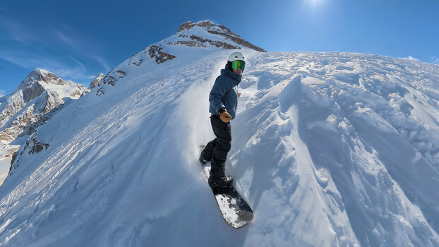 Snowboarder riding down steep snowy mountain slope in bright sunlight