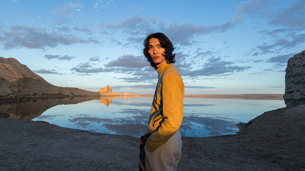 Man in golden hour light beside reflective lake with mountains and rock formations