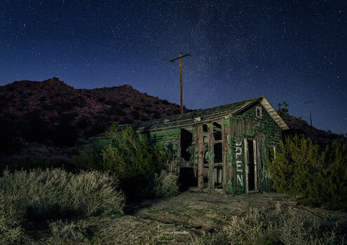 Abandoned green wooden house at night beneath starry sky with desert hills