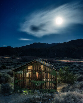 Abandoned wooden cabin illuminated by moonlight in desert landscape with mountains