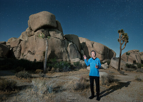 Portrait of man in blue shirt standing in desert landscape with massive granite boulders and Joshua tree under starry night sky