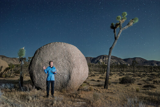 Person in blue shirt standing beside massive spherical boulder under starry night sky in desert landscape